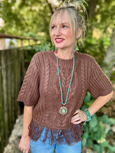 A woman wearing a brown short-sleeve cable-knit sweater with fringe trim, styled with blue jeans, turquoise jewelry, and a long turquoise beaded necklace, standing outdoors in front of a wooden fence and green foliage.