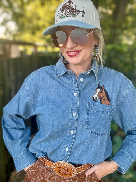 A woman wearing a light blue denim long-sleeve button-up shirt with a embroidered brown horse on the chest pocket, paired with a white and blue western-style baseball cap, aviator sunglasses, and a brown western belt.