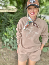Woman wearing a beige sweatshirt with a graphic design, shorts, and a cap with a scarf in an outdoor setting.