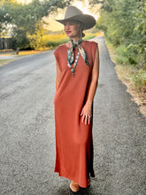 Woman in a rust-colored dress and wide-brimmed hat standing on a road with greenery around.