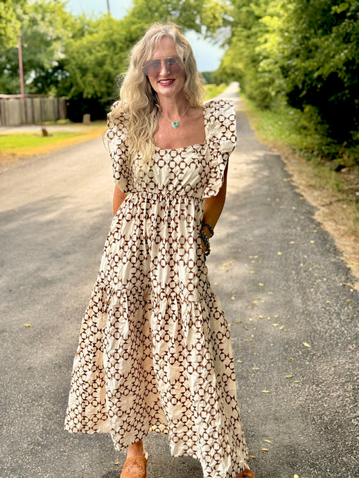 A woman wearing a bohemian-style tiered maxi dress with a brown and cream geometric pattern, standing on an outdoor country road.