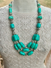 A multi-strand statement necklace featuring vibrant turquoise beads and textured silver-toned metal spacers, displayed on a cream-colored lace-knit sweater mannequin.