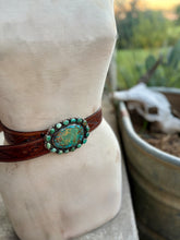 A brown leather wrap bracelet with a large oval turquoise and green stone centerpiece, displayed on a cream fabric mannequin stand with an outdoor blurred background.