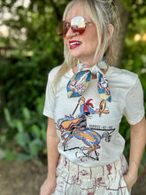 Woman wearing a graphic t-shirt with a horse and rooster design, sunglasses, and a scarf outdoors.
