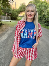 Woman wearing a blue t-shirt with 'America Mama' text and red checkered shirt and shorts on a road.