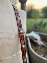 A close-up of a brown leather strap with embossed floral patterns and silver-toned conchos, draped over a beige fabric mannequin torso with an outdoor blurred background.