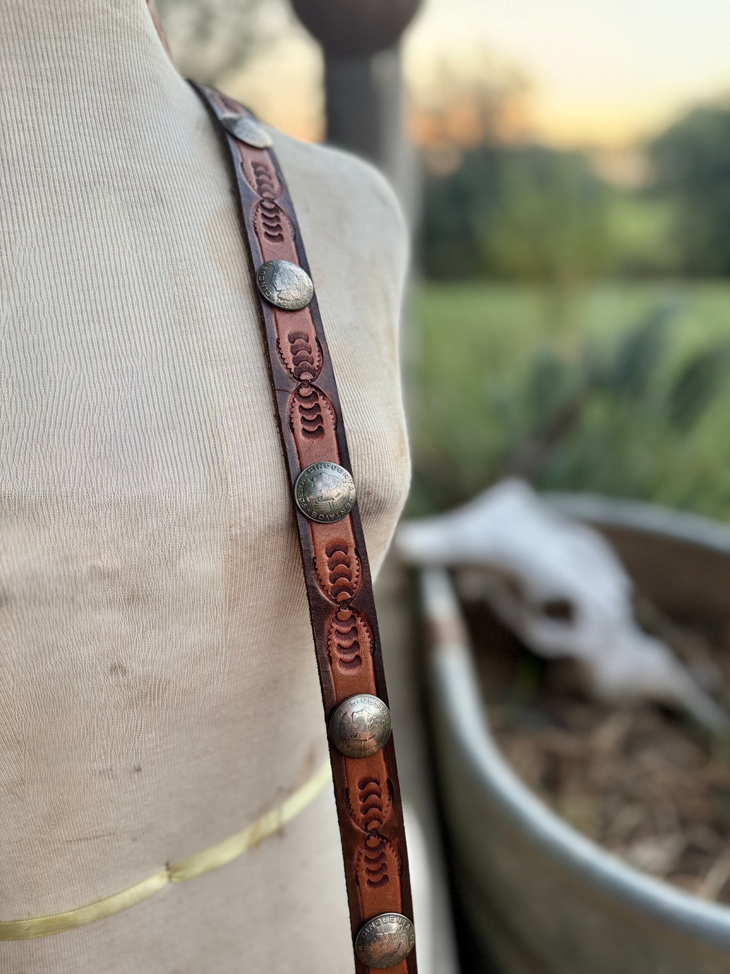 A close-up of a brown leather strap with embossed floral patterns and silver-toned conchos, draped over a beige fabric mannequin torso with an outdoor blurred background.