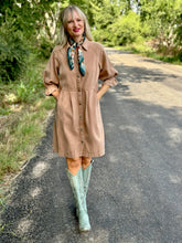 Woman in a brown dress and green boots standing on a path with trees in the background