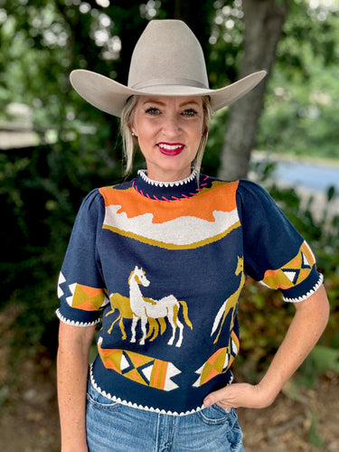 Woman wearing a navy sweater with horse designs and a cowboy hat outdoors.