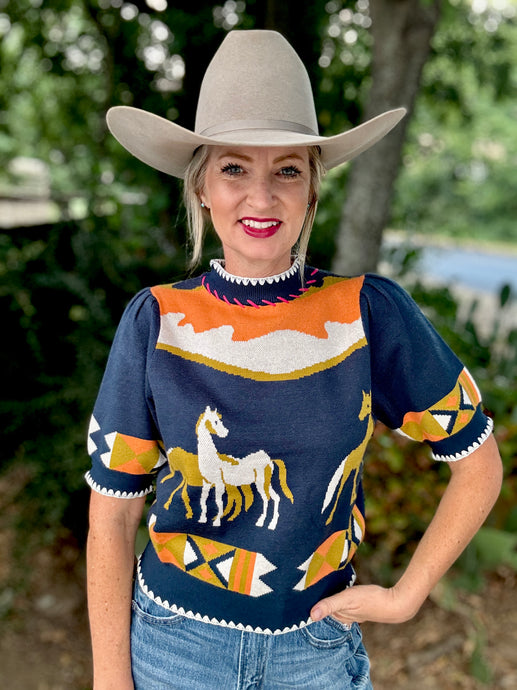 Woman wearing a navy sweater with horse designs and a cowboy hat outdoors.