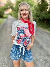 Woman wearing a white t-shirt with an American flag design, denim shorts, and a red bandana outdoors.