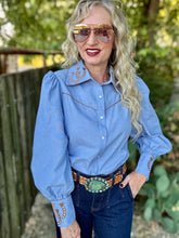 A woman wearing a light blue western-style long-sleeve button-up shirt with decorative embroidery and contrast stitching on the chest and cuffs, paired with dark jeans and a large western belt buckle, standing outdoors in front of a wooden fence and green foliage.