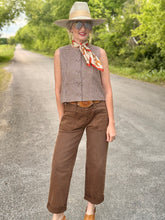 Woman in a sleeveless checkered top and brown pants standing on a road with greenery in the background.