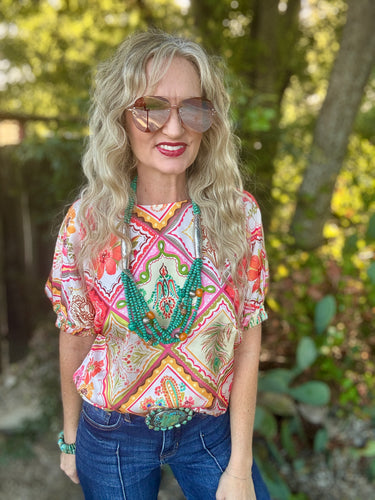 A woman with long blonde wavy hair and sunglasses wearing a colorful bohemian-style patterned blouse with blue jeans, standing outdoors against a blurred green background.