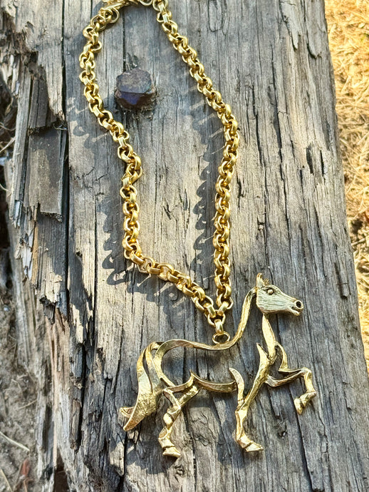A gold horse pendant necklace on a matching chain, displayed on a weathered wooden log outdoors.