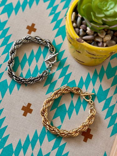 Two braided bracelets, one gold and one silver, on a patterned surface with a potted plant.
