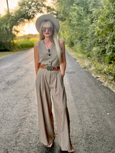 Woman in a beige sleeveless top and wide-leg pants standing on a road with trees in the background.