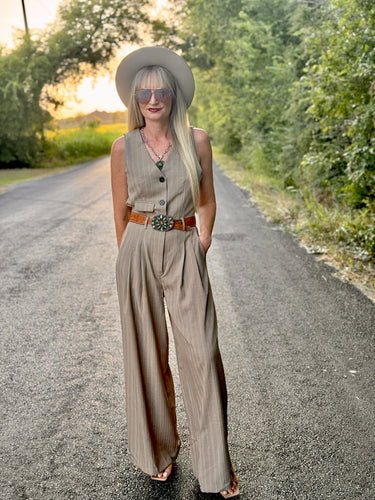 Woman in a beige sleeveless top and wide-leg pants standing on a road with trees in the background.