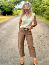 A woman stands on a paved country road wearing a cream sleeveless knit top, brown cropped pants, and brown suede mules, with trees and grass in the background.