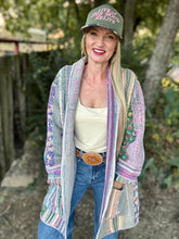 Woman wearing a colorful cardigan, white tank top, jeans, and a green cap with text outdoors.