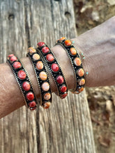 A close-up of a person's wrist wearing a stack of ornate black metal bangles decorated with oval red, orange, and pink stones, photographed outdoors against a weathered wooden surface.