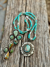 A turquoise and silver jewelry set displayed on a weathered wooden surface, featuring a long beaded necklace and two matching rings with green and brown stones.
