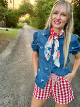 Woman wearing a denim shirt with star patterns, a red, white, and blue scarf, and red and white checkered shorts on a road.