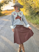 Woman in a gray sweater with a horse design and brown skirt standing on a road.