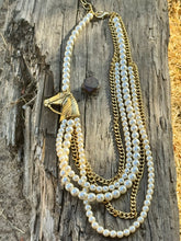 A multi-strand necklace featuring white faux-pearl strands and gold-tone chains, with a horse-head charm, displayed on a weathered wooden log outdoors.