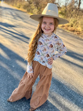 Young girl in a patterned shirt and wide-leg pants with a hat standing on a road.