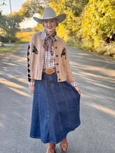 Person wearing a cowboy hat, patterned shirt, beige cardigan, blue denim skirt, and brown boots on a road with trees in the background.