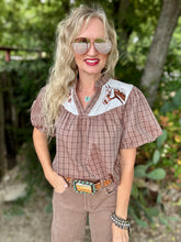 A woman wearing a brown and beige plaid short-sleeve blouse with a white collar featuring embroidered brown horse designs, paired with brown pants and a western-style belt, standing outdoors in front of a blurred green background.