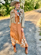 Woman in a long orange dress with white patterns, beige jacket, and brown boots standing on a road.