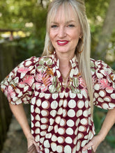 A smiling blonde woman wearing a short-sleeve blouse with a burgundy base and a cream polka dot pattern, accented with pink and green floral details on the chest and sleeves, set against a blurred outdoor background.
