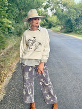 Woman wearing a sweater with a horse design and wide-brimmed hat on a road with trees in the background