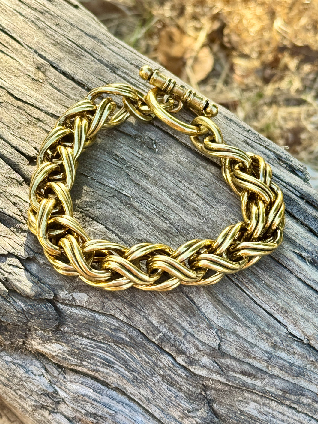 A thick gold-toned braided chain bracelet with a toggle clasp, displayed on a weathered gray wooden log with a blurred outdoor background.