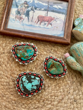 Three western-style turquoise and black stone buckles with red bead borders and silver-toned metal settings, arranged on a woven beige surface with a small framed western painting and decorative cactus figurines nearby.
