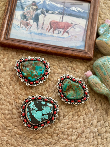 Three western-style turquoise and black stone buckles with red bead borders and silver-toned metal settings, arranged on a woven beige surface with a small framed western painting and decorative cactus figurines nearby.