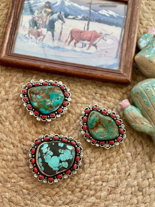 Three western-style turquoise and black stone buckles with red bead borders and silver-toned metal settings, arranged on a woven beige surface with a small framed western painting and decorative cactus figurines nearby.