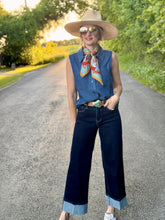 Woman in a denim tank top and wide-leg jeans standing on a road with trees in the background