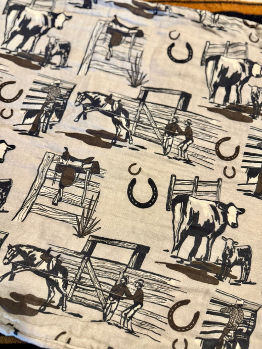 A close-up of a western-themed fabric featuring black illustrations of cows, horses, riders, ladders, and horseshoes on a warm off-white background.