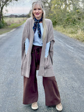 Woman standing on a road wearing a beige cardigan, white shirt, brown wide-leg pants, and a patterned scarf.