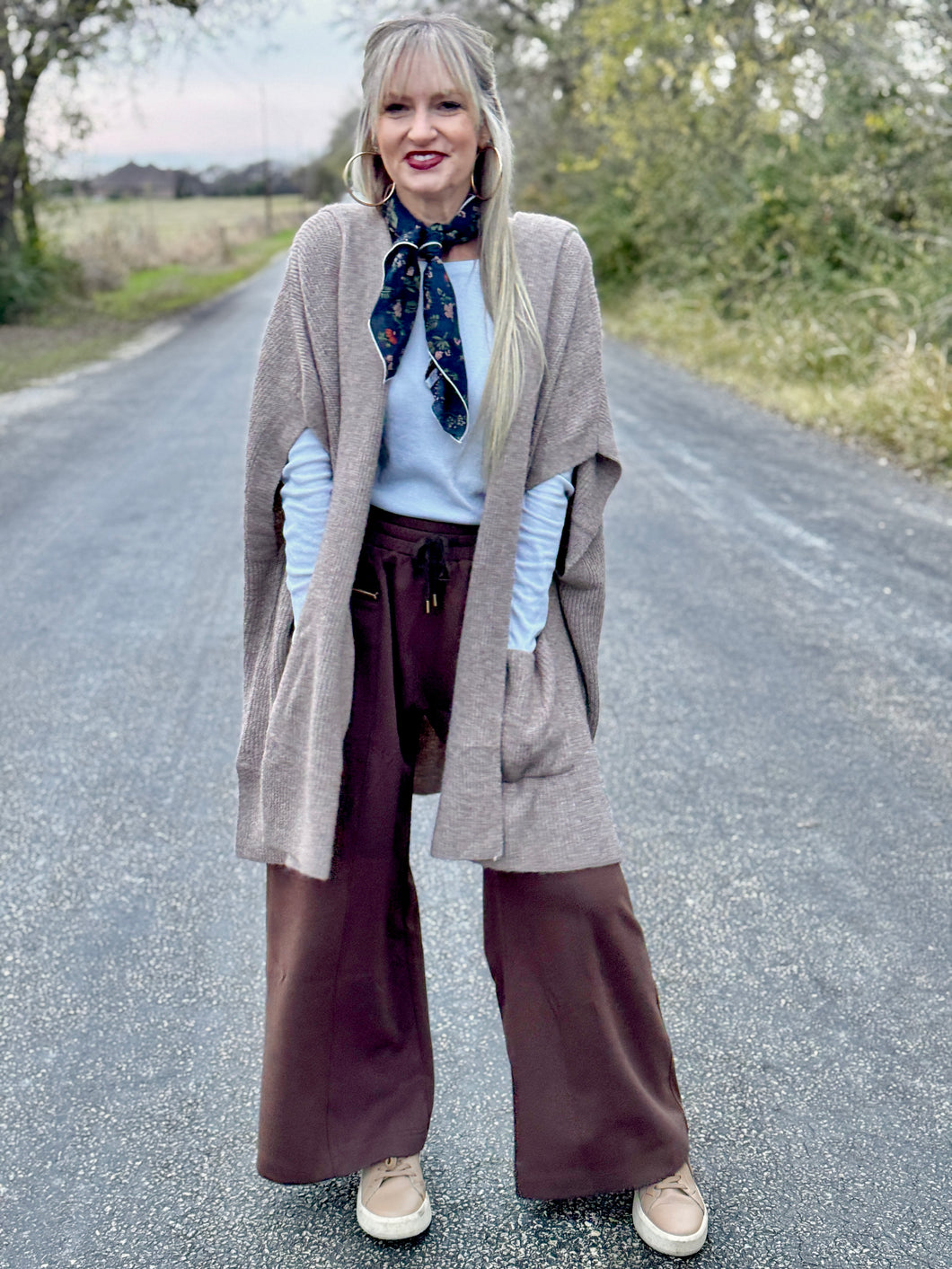Woman standing on a road wearing a beige cardigan, white shirt, brown wide-leg pants, and a patterned scarf.