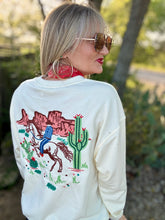 Woman wearing a white sweatshirt with a desert-themed design outdoors.
