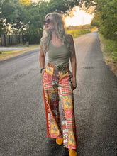 A woman stands on a paved country road at sunset wearing a sleeveless olive green top and highly patterned patchwork wide-leg pants with orange side panels, yellow gingham sections, and brown floral motifs. She is wearing sunglasses and yellow shoes.
