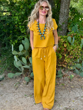 A woman wearing a mustard yellow short-sleeve top and matching pants with a drawstring waist, standing outdoors in front of a garden with cacti and trees.
