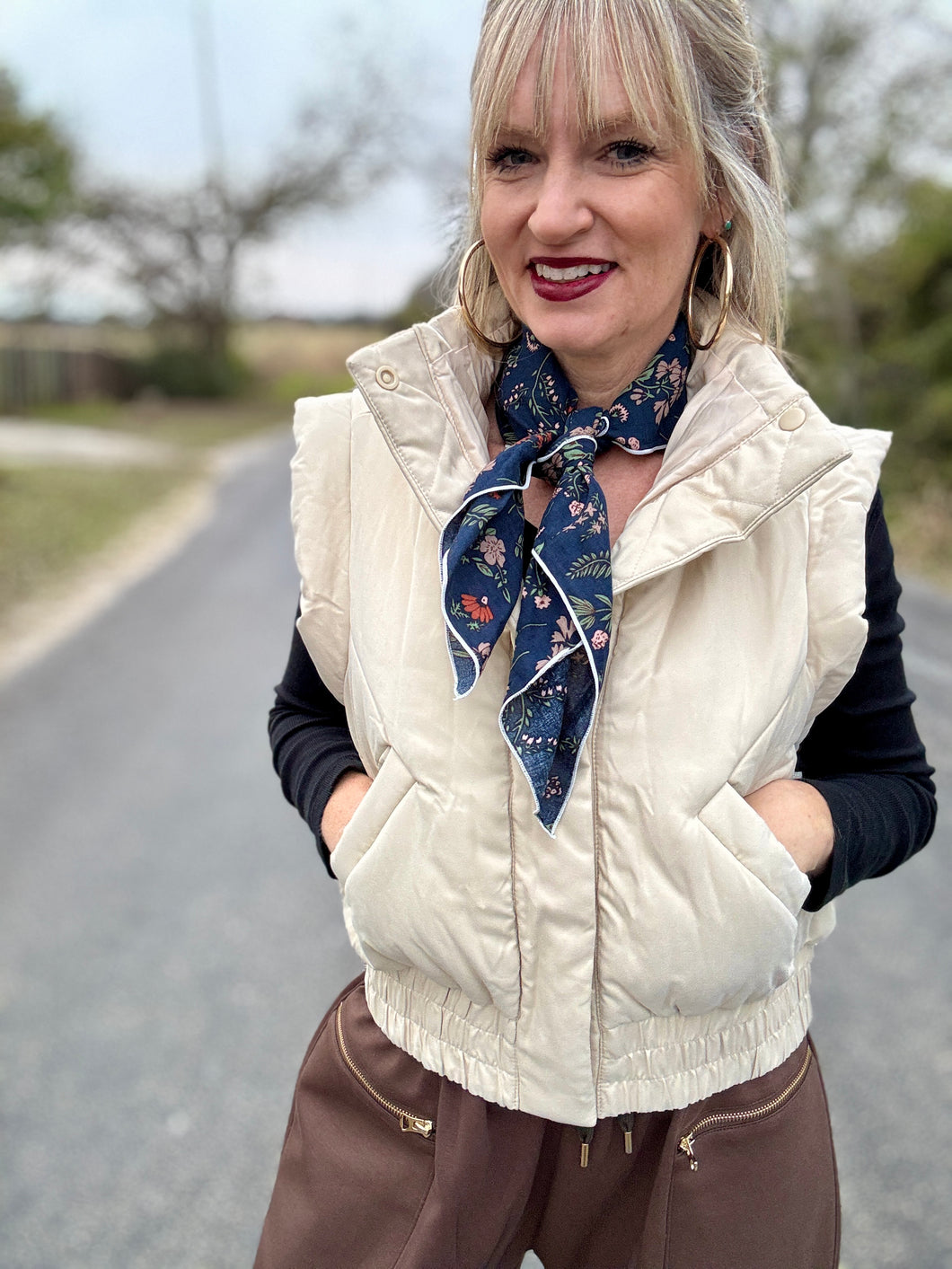 Woman wearing a white puffer vest and brown pants with a blue floral scarf around her neck, standing on a road.