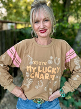 A woman wearing a tan long-sleeve sweatshirt with pink and white western-themed graphics and the text 'You're MY LUCKY CHARM', paired with blue jeans and a large turquoise belt buckle, standing outdoors with a blurred green background.