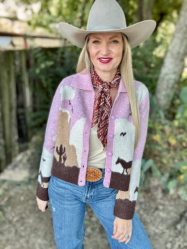 Woman wearing a patterned cardigan, jeans, and a cowboy hat outdoors.