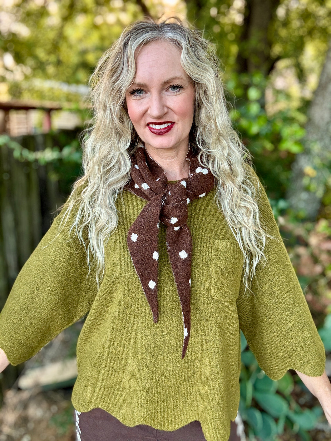 A woman with long blonde wavy hair wearing a textured olive green top and a brown neck scarf with white polka dots, standing outdoors in front of a blurred green background.